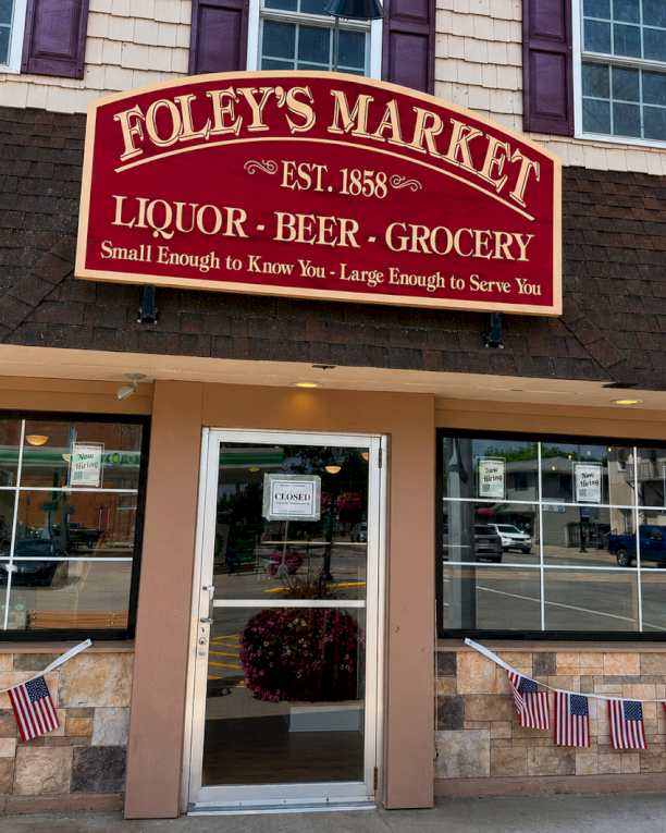 Foley&rsquo;s Market sign outside a store labeled liquor, beer, and grocery, with flags along the entrance.